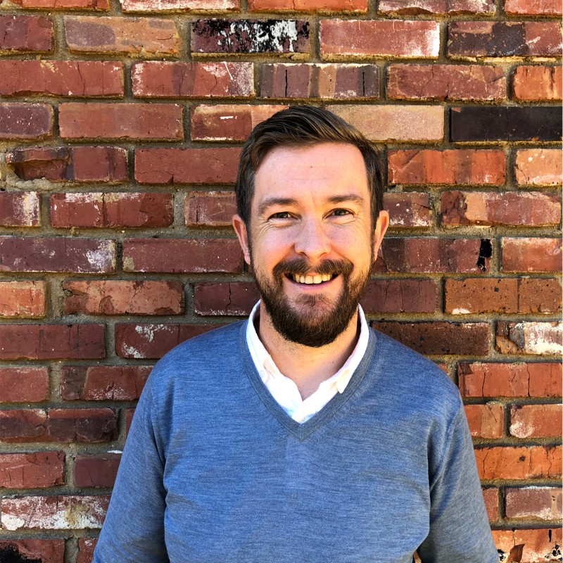 Headshot of Jon wearing a blue sweater with a brick wall behind him.